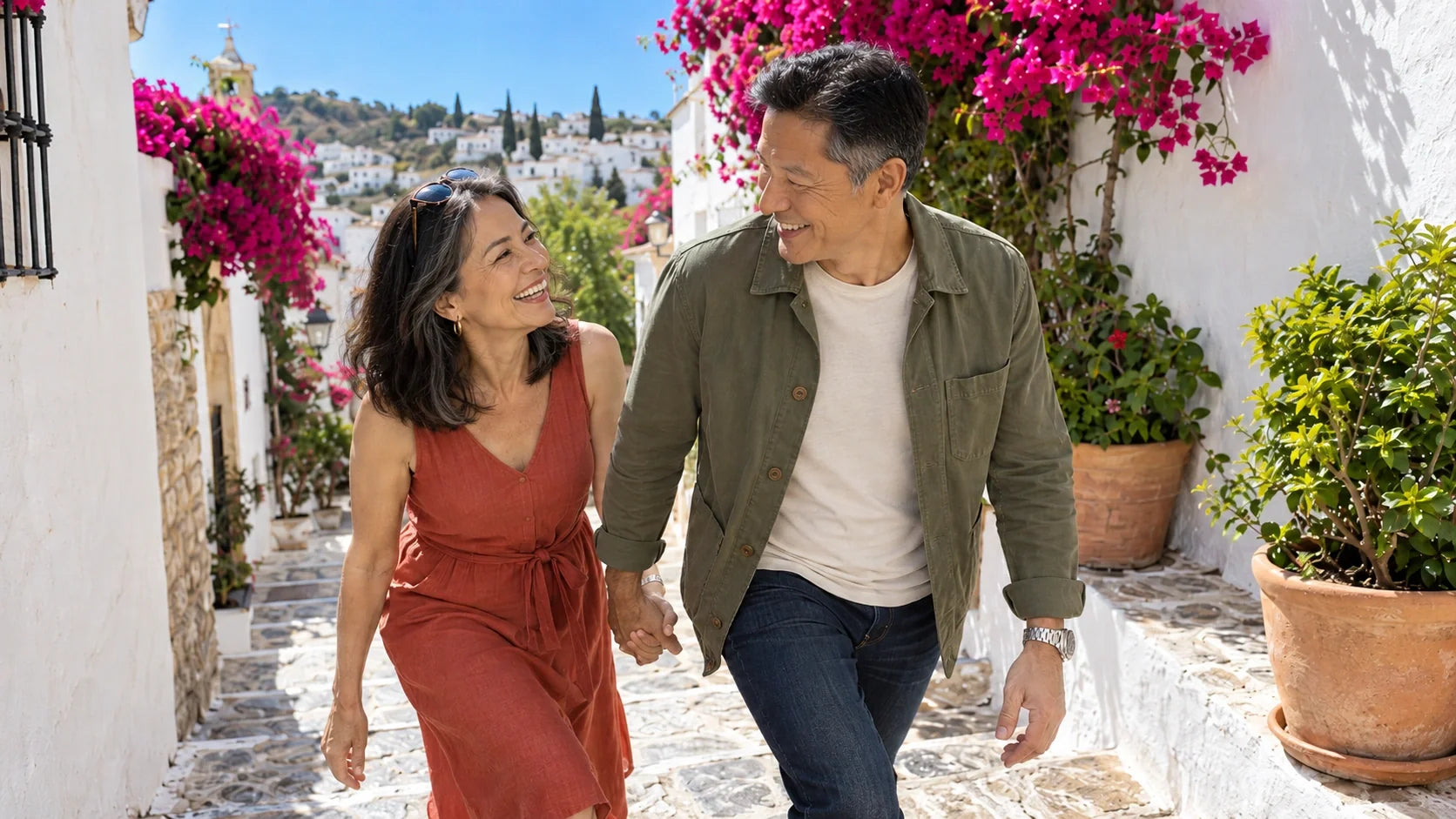 A couple in their early 60s laughing as he leads her by the hand up a sunny whitewashed village lane